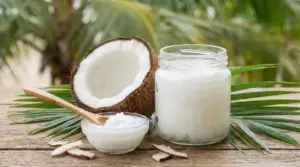 Vasetto di olio di cocco con mezzo cocco e cucchiaio su tavolo di legno, con foglie di palma sullo sfondo