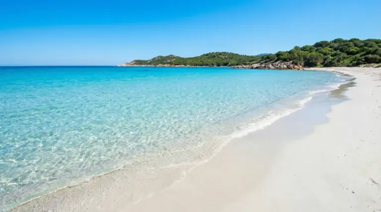 Spiaggia di sabbia bianca con mare turchese e acqua cristallina, costa verde sullo sfondo in una baia tranquilla